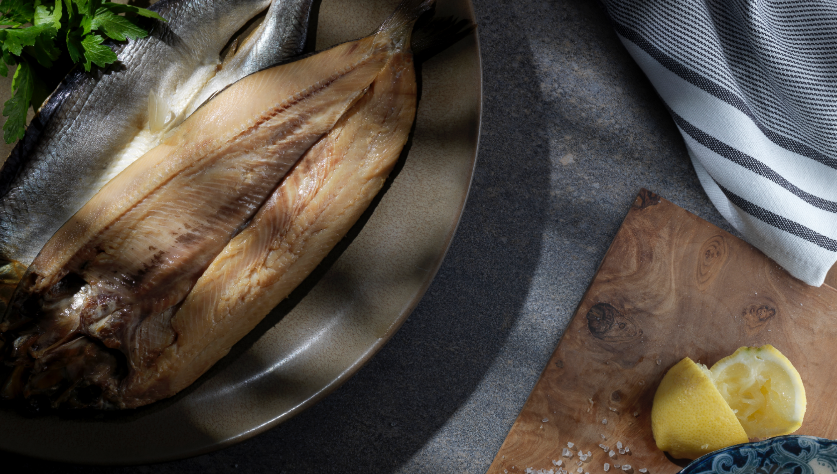 Two smoked Mallaig kippers resting on a brown dinner plate, with a fresh lemon nearby for seasoning.